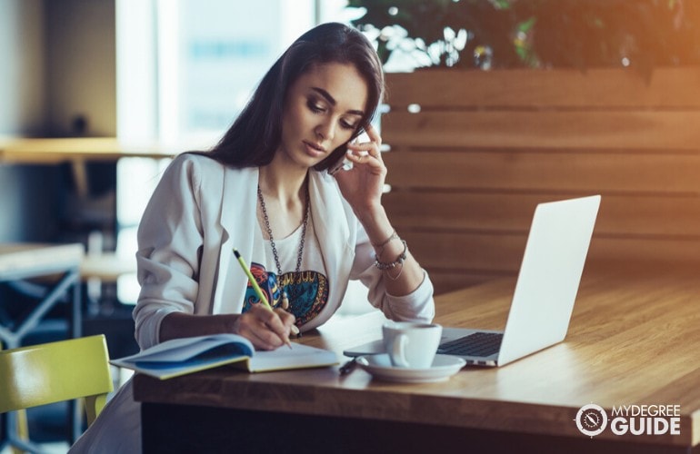 marketing expert talking on the phone while writing on her notebook in a cafe