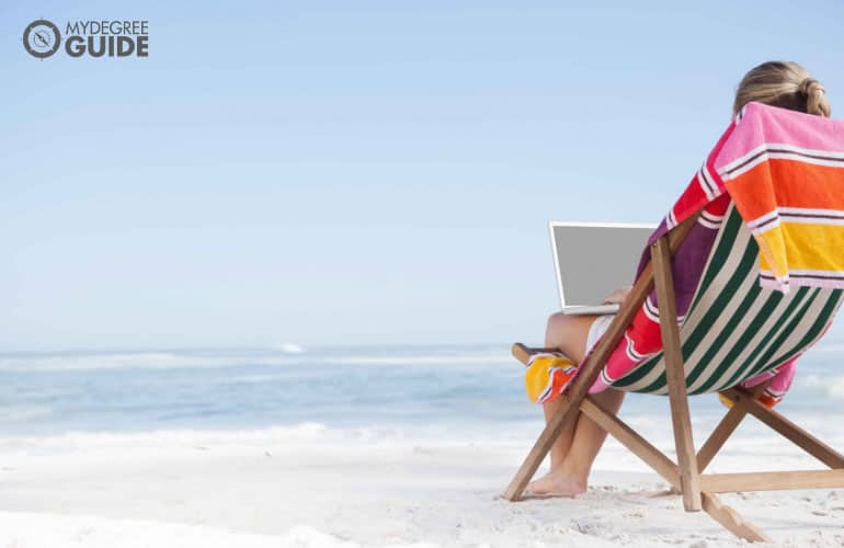 female sitting on a beach looking at their laptop